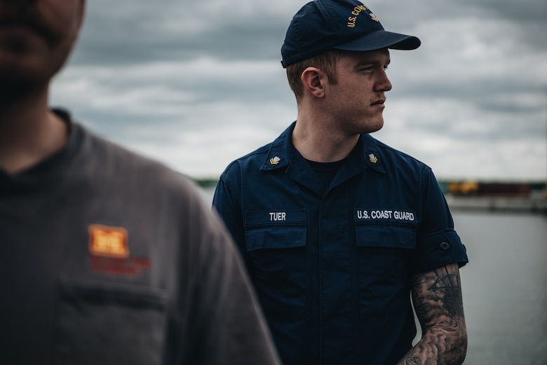 A man in a navy blue U.S. Coast Guard uniform and hat looks off to his left. Another figure is out of focus in the foreground.