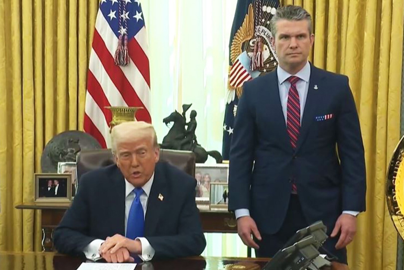 A man sits while one stands behind a desk in an office.