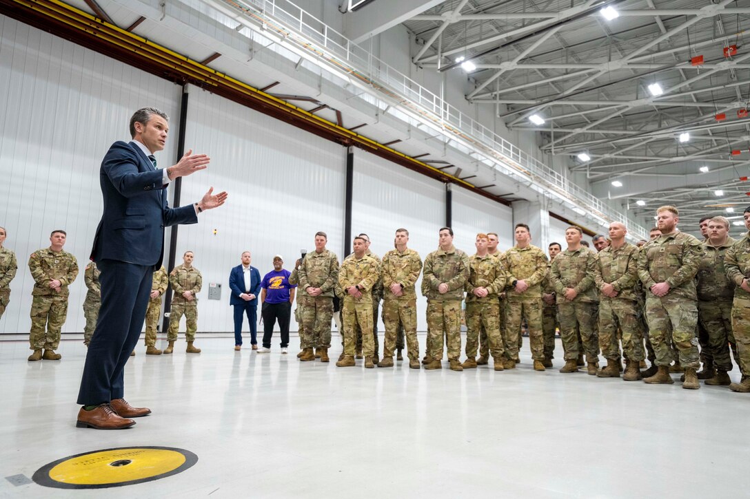 Defense Secretary Pete Hegseth uses his hands while talking with troops in a semi-circle formation in a large hangar.