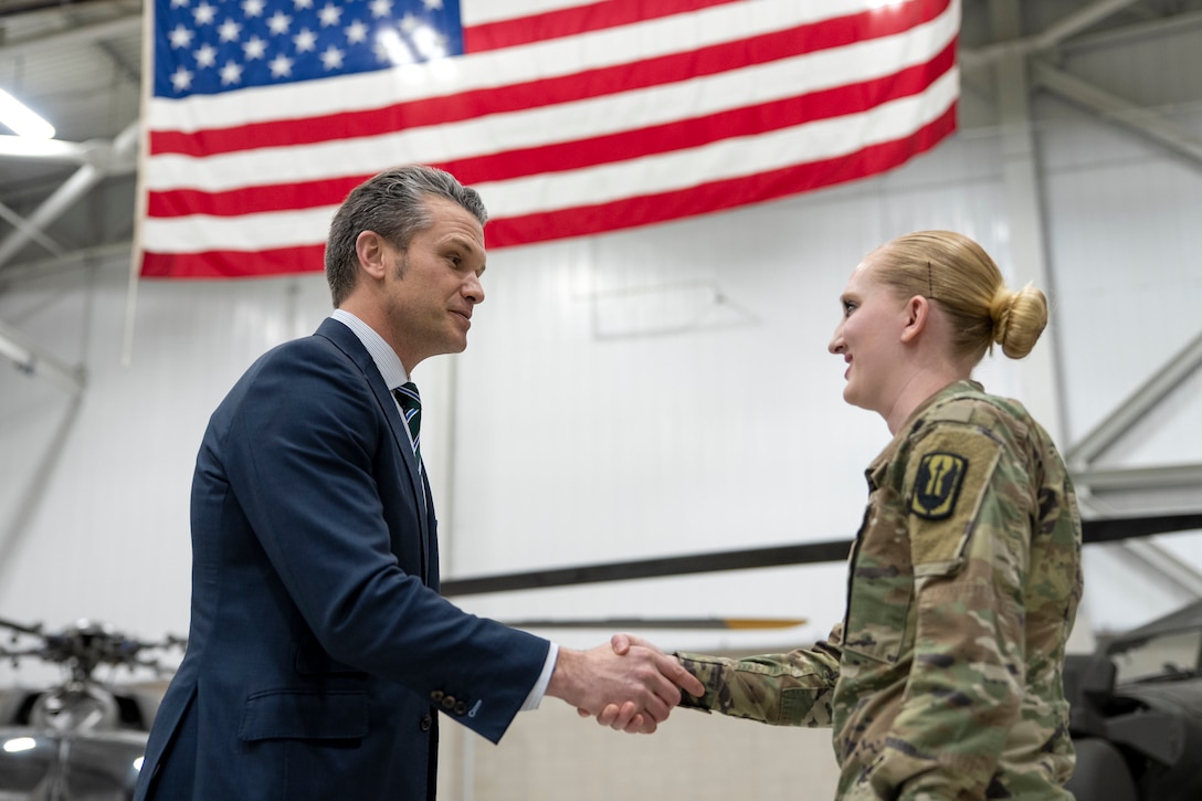 Defense Secretary Pete Hegseth shakes hands with a service member in a hangar with the American flag overhead.