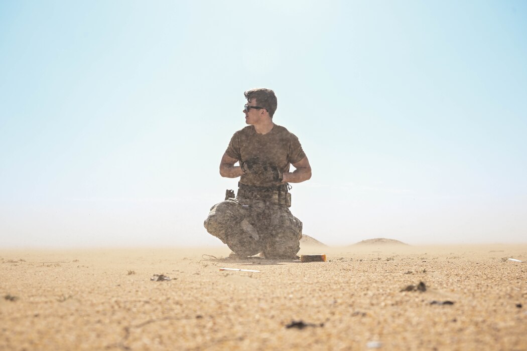 A service member kneels in the sand as he takes off his gloves.
