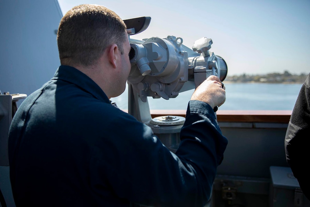 A close-up of a sailor looking through large binoculars aboard a ship at sea.