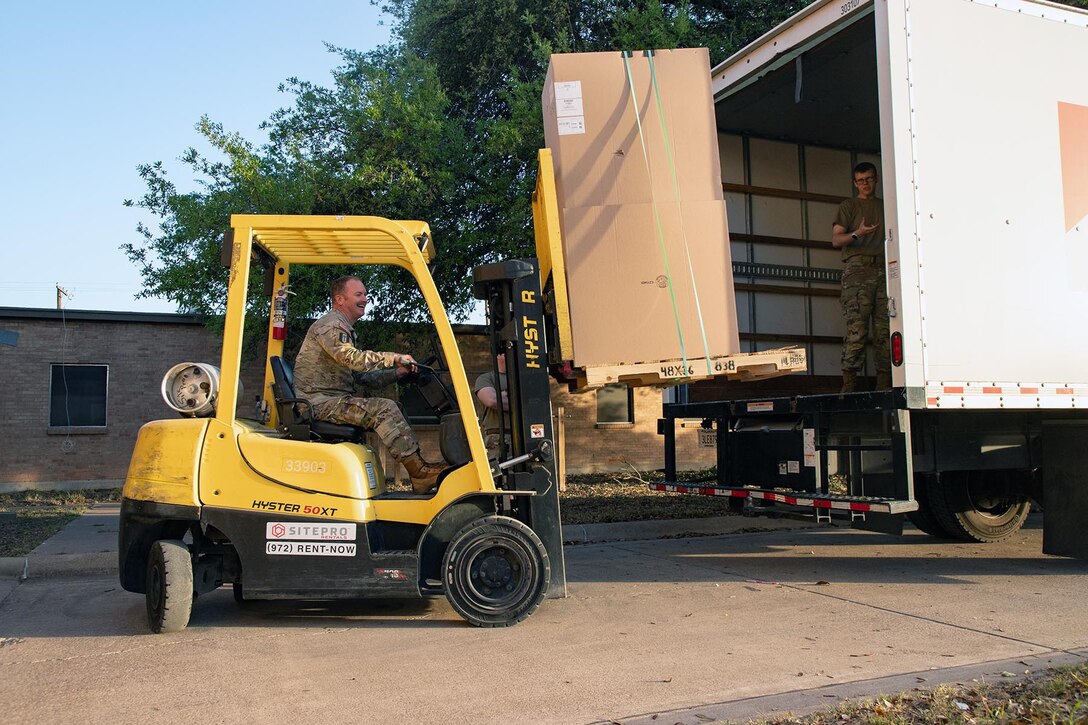 A soldier drives construction equipment to move a large box from a moving truck.