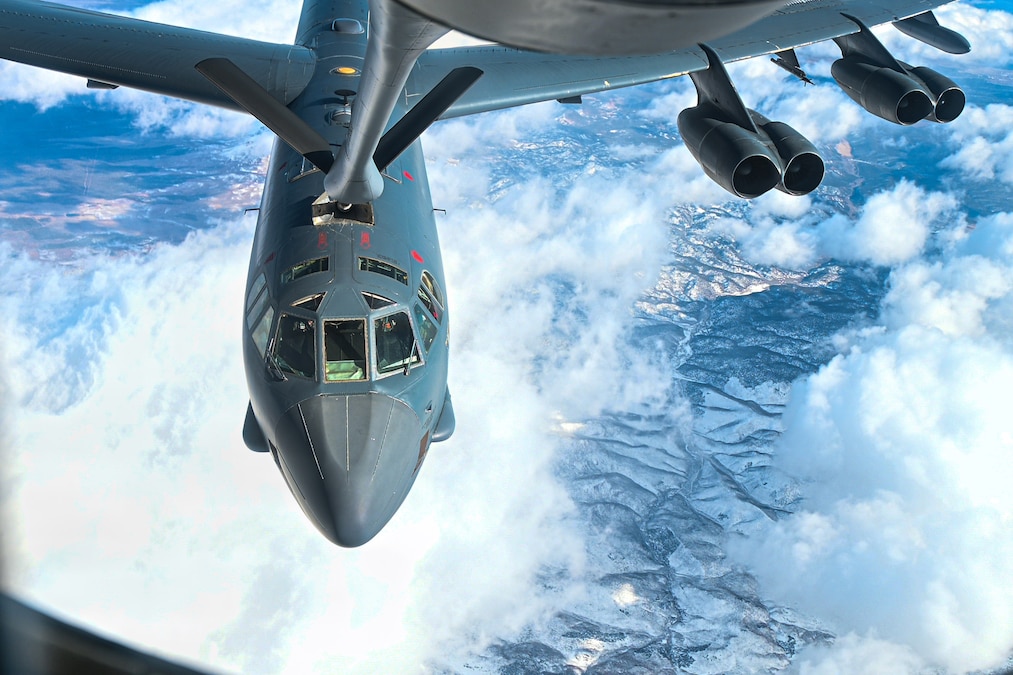 A plane maneuvers under another plane high above clouds and mountains.