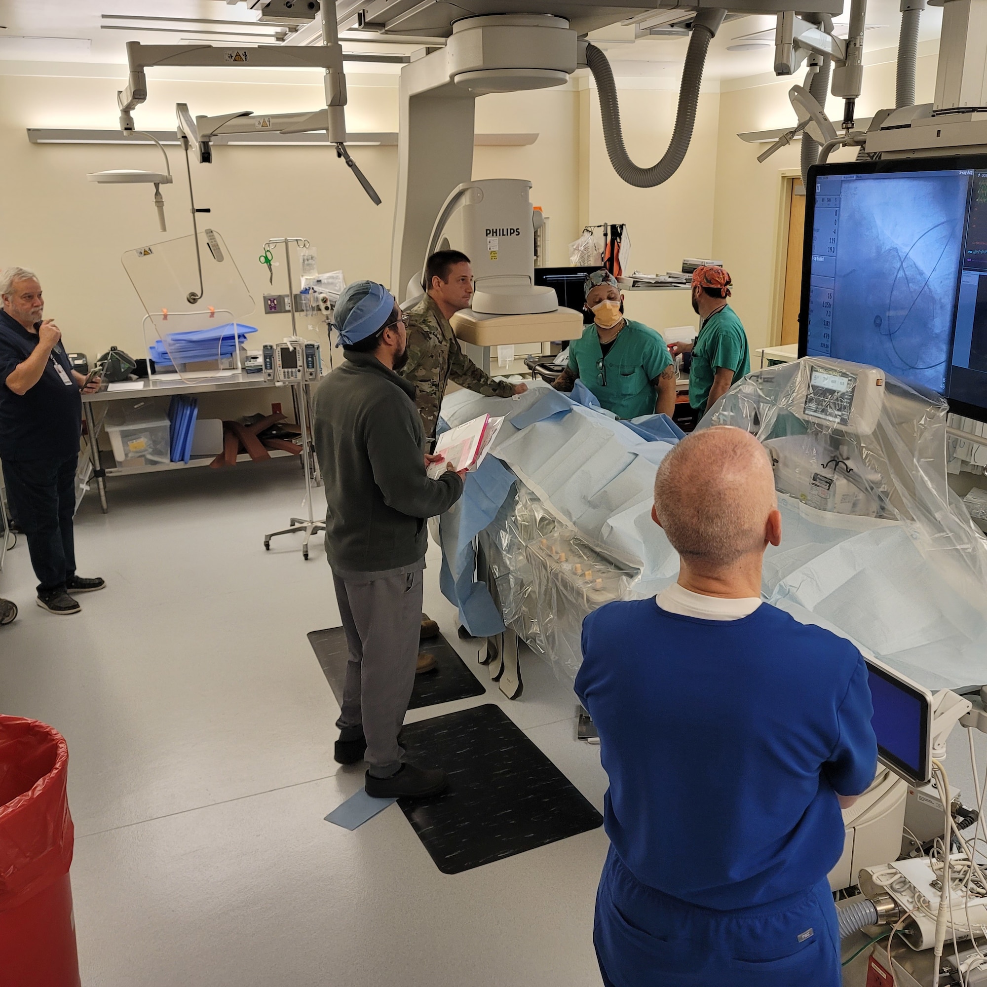 Five individuals from right to left, one man in royal blue scrubs, a man in a bullet grey jacket wearing grey pants, one in camouflage, and two men in turquoise scrubs surround an operating table.