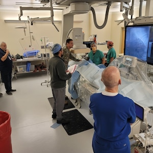 Five individuals from right to left, one man in royal blue scrubs, a man in a bullet grey jacket wearing grey pants, one in camouflage, and two men in turquoise scrubs surround an operating table.