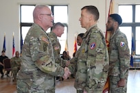 An Army soldier in Army combat uniform is shaking hands with another similarly dressed Army soldier in a large room during a ceremony