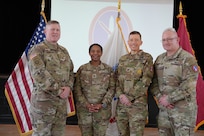 Four Army Soldiers who are wearing Army combat uniforms are standing in a line while posing for a picture with their hands together in front of their waists. Behind them are three flags and a projection of the MDW logo on a screen in the background.
