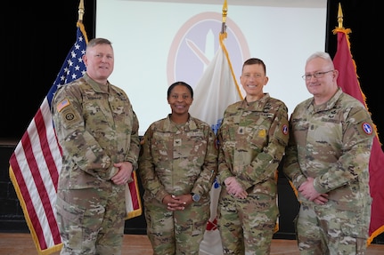 Four Army Soldiers who are wearing Army combat uniforms are standing in a line while posing for a picture with their hands together in front of their waists. Behind them are three flags and a projection of the MDW logo on a screen in the background.