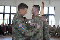 Two Army Soldiers wearing Army combat uniforms are facing each other during a ceremony and the Soldier on the left is changing the patch on the other Soldier's shoulder.