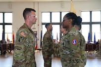 Two Army Soldiers wearing the Army combat uniform are facing each other in a room during a ceremony.