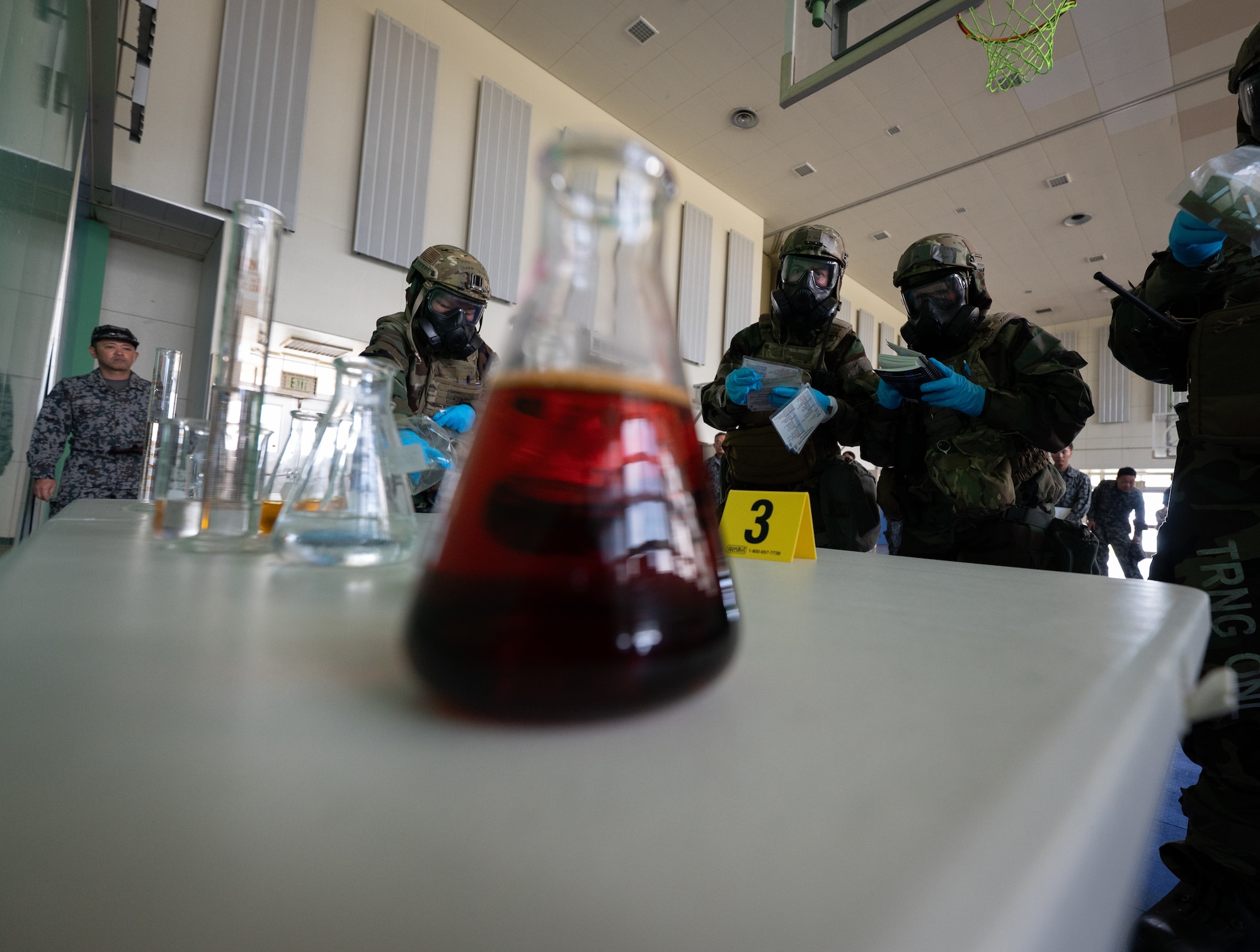 Japanese Air Self-Defense Force members watch U.S. service members in chem gear test chemicals.