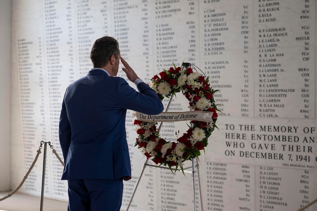 Defense Secretary Pete Hegseth salutes a wreath on a stand in front of a white wall with names inscribed on it.