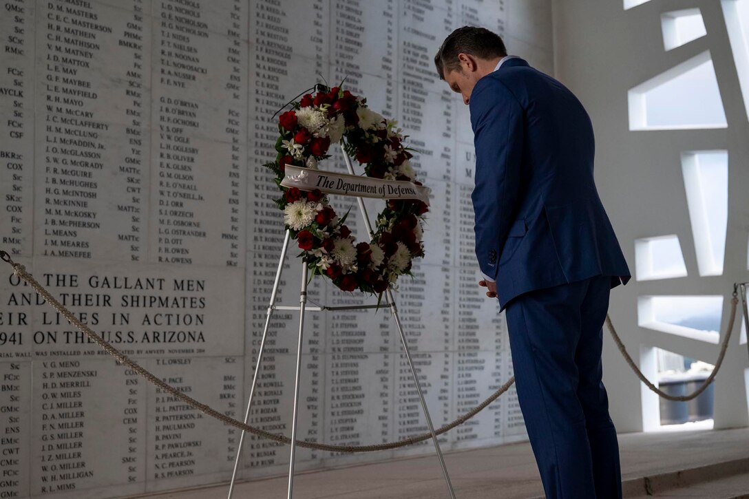 Defense Secretary Pete Hegseth bows his head in front of a wreath on a stand in front of a white wall with names inscribed on it.