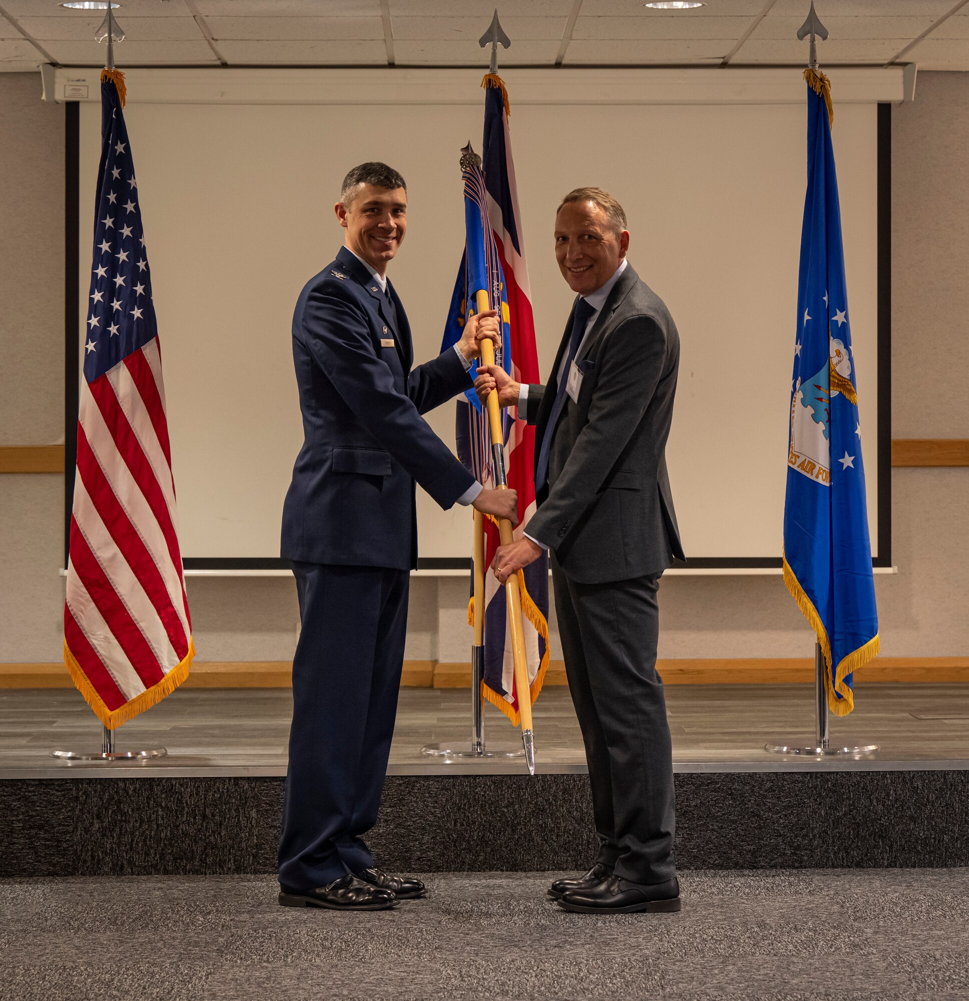 U.S. Air Force Col. Aaron Rivers, left, 100th Maintenance Group commander, presents a guidon to Julian Long, Electronic Management Services managing director, during the honorary commanders’ induction ceremony at RAF Mildenhall, England, March 21, 2025.