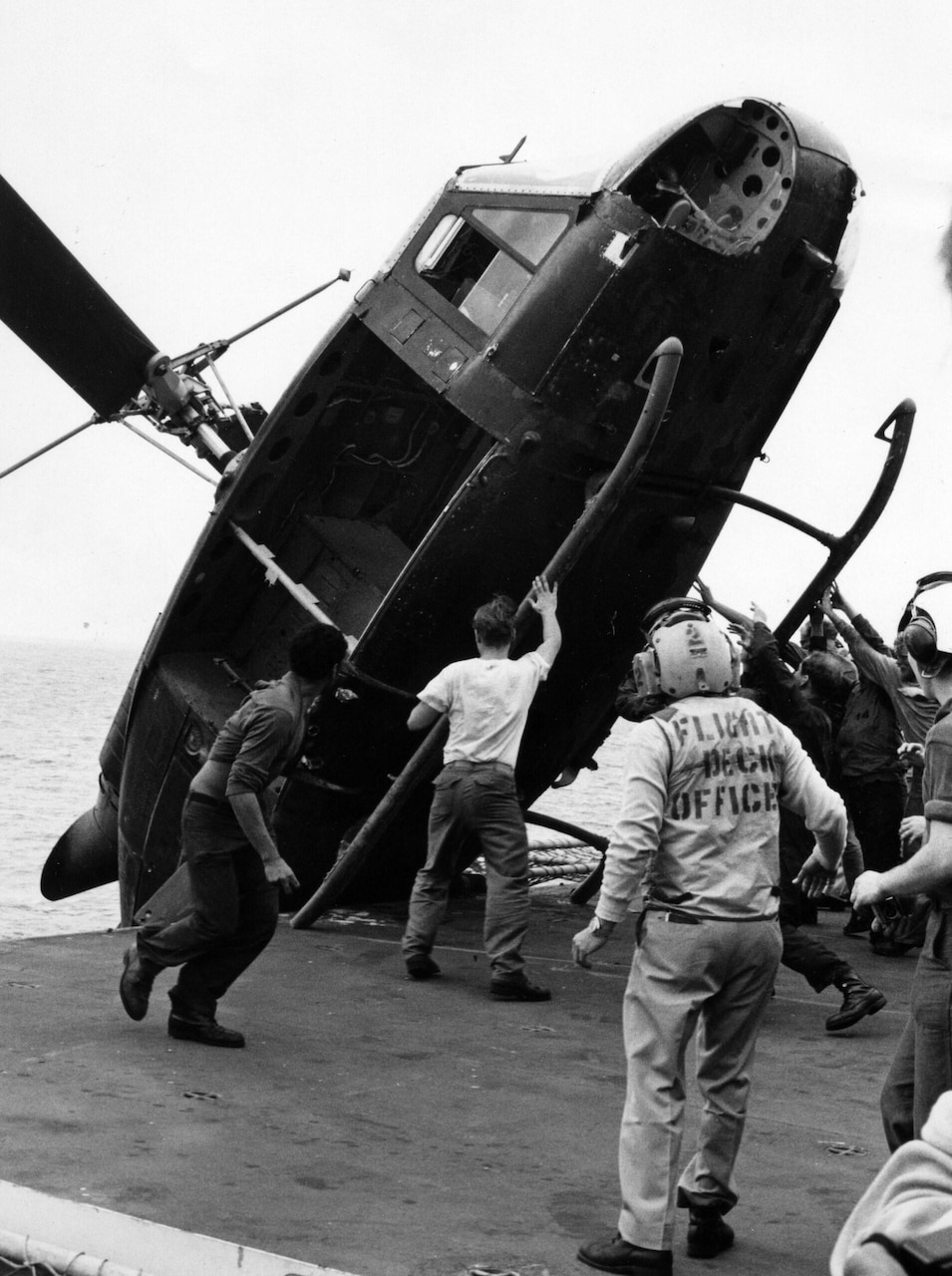 A group of men pushes a helicopter off the side of a ship.
