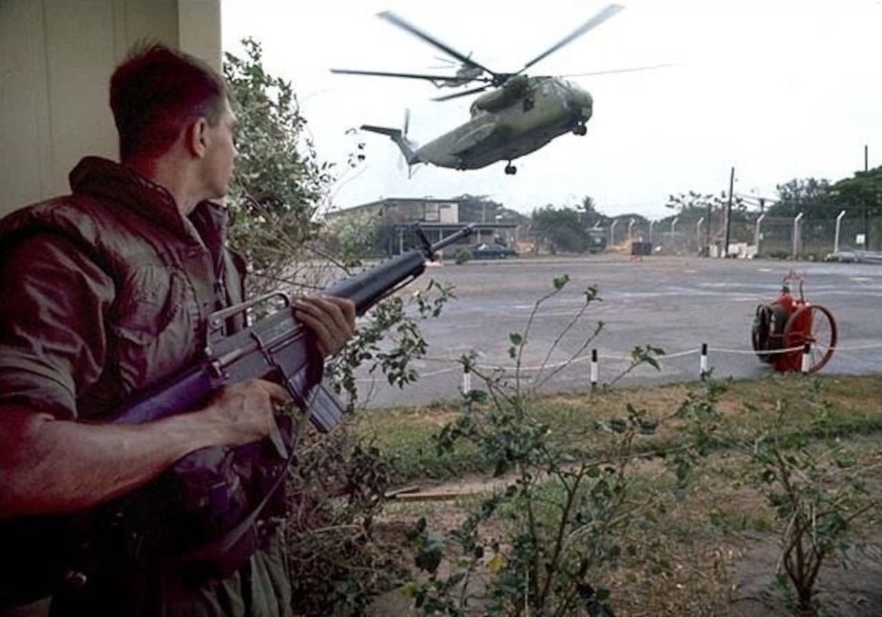 A man holds a gun and watches as a helicopter lands.