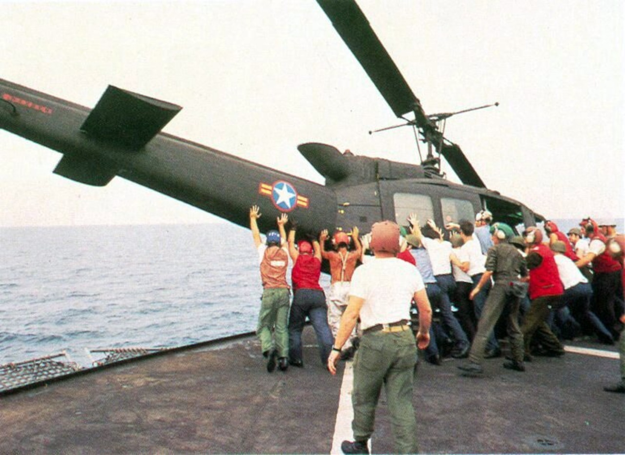 More than a dozen men in uniforms push a helicopter off the side of a ship.