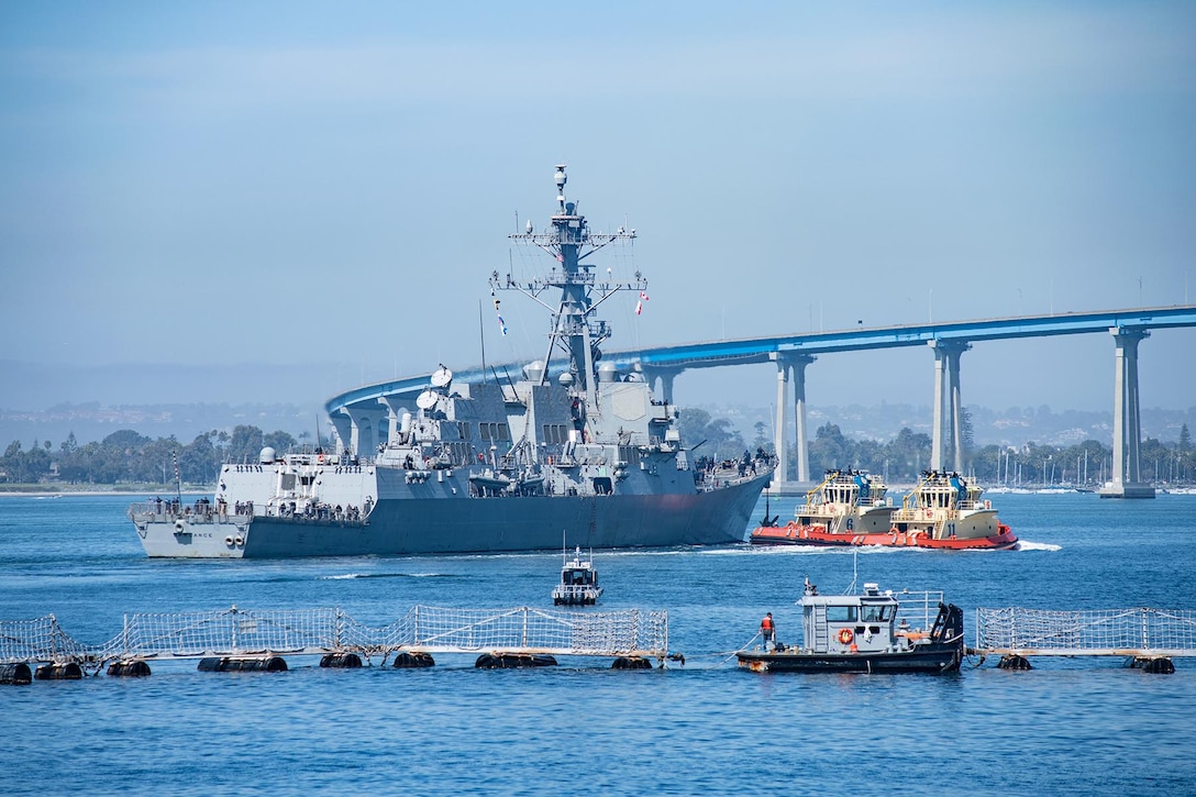 A ship travels near smaller vessels, a raised bridge and shoreline.