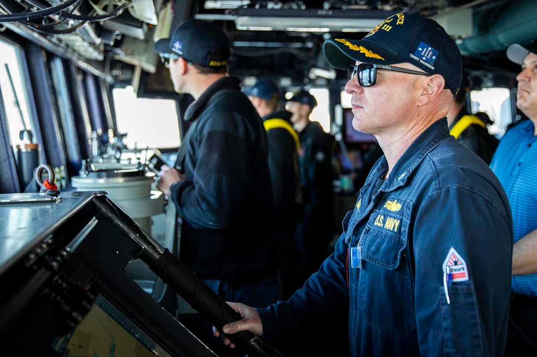 A Navy commander stands looking out the windows of a ship alongside other sailors doing the same.