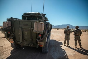 Two people wearing camouflage uniforms stand alongside a large military vehicle with a mountain range in the background.