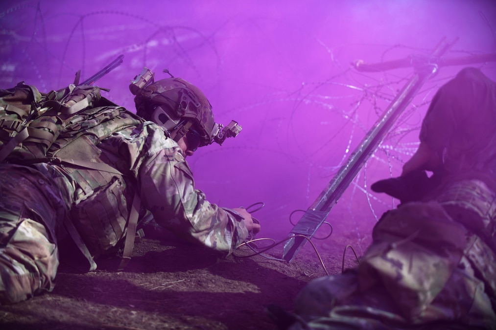 Soldiers in tactical gear lay in the dirt near barbed wire surrounded by clouds of purple smoke.