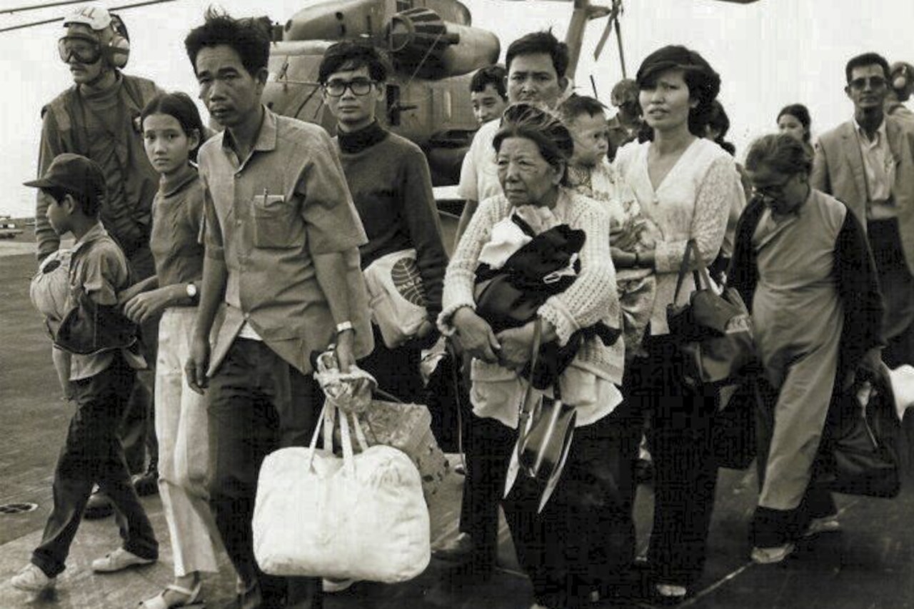 A large group of people holding their belongings as they arrive aboard a Navy vessel. A helicopter is in the background.