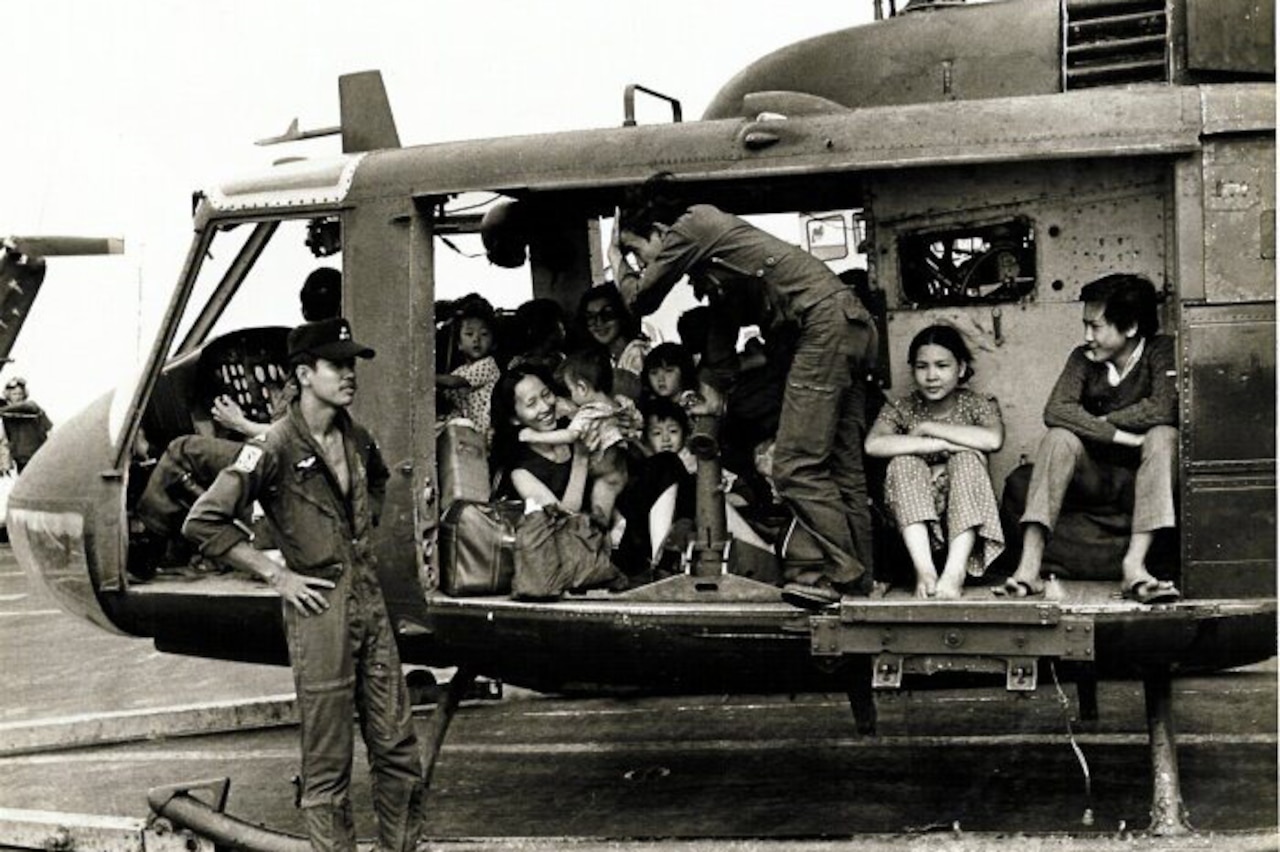 A helicopter sits on the ground with a group of adults and children inside while a man in a military uniform stands beside the aircraft.