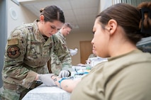 A 5th Medical Group Airman sticks a needle into another Airman's arm for IV training.