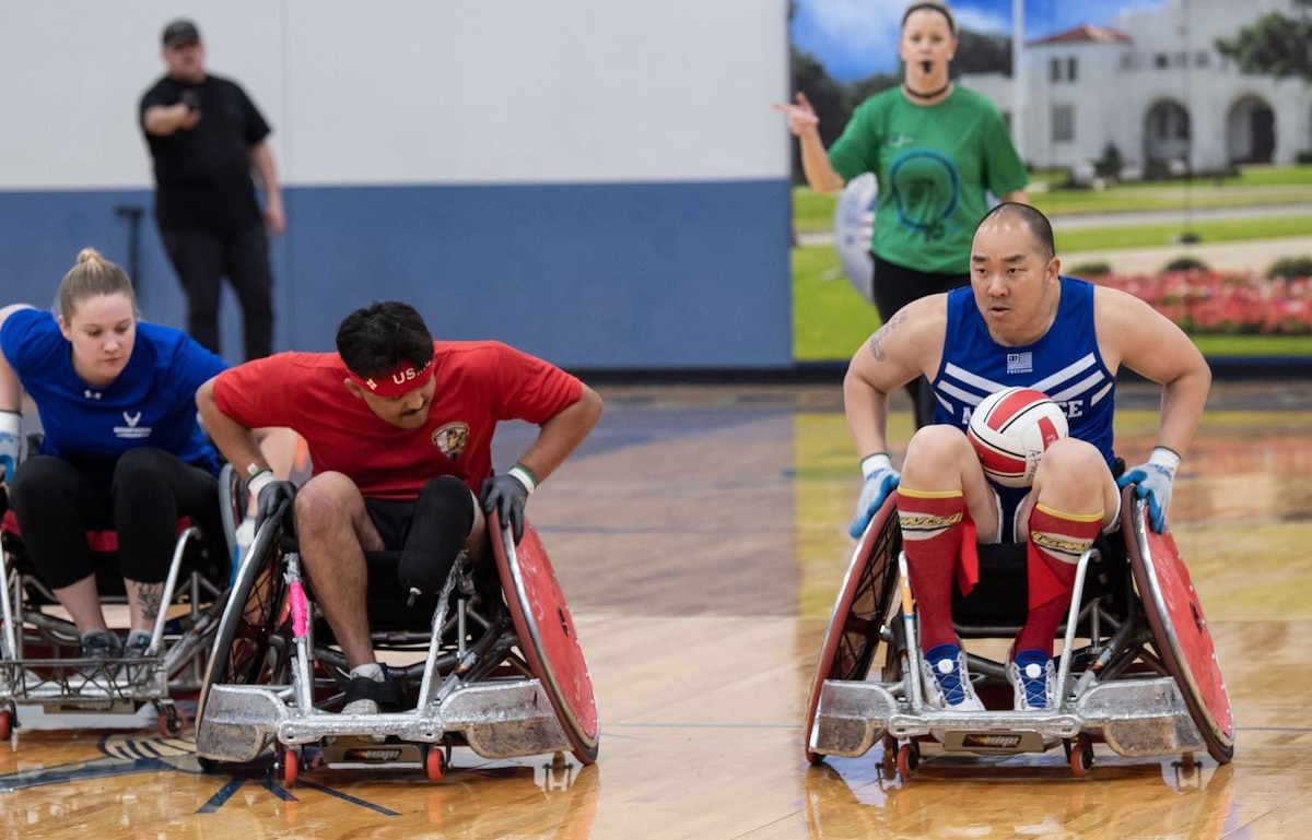 Lin in a wheelchair at right has the ball while a marine corps athlete tries to catch him. Another Air Force athlete is just behind the Marine.