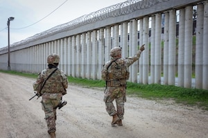 Two people wearing camouflage uniforms walk down a dirt road as the person on the right points to a large wall with concrete pillars and barbed wire at the top.