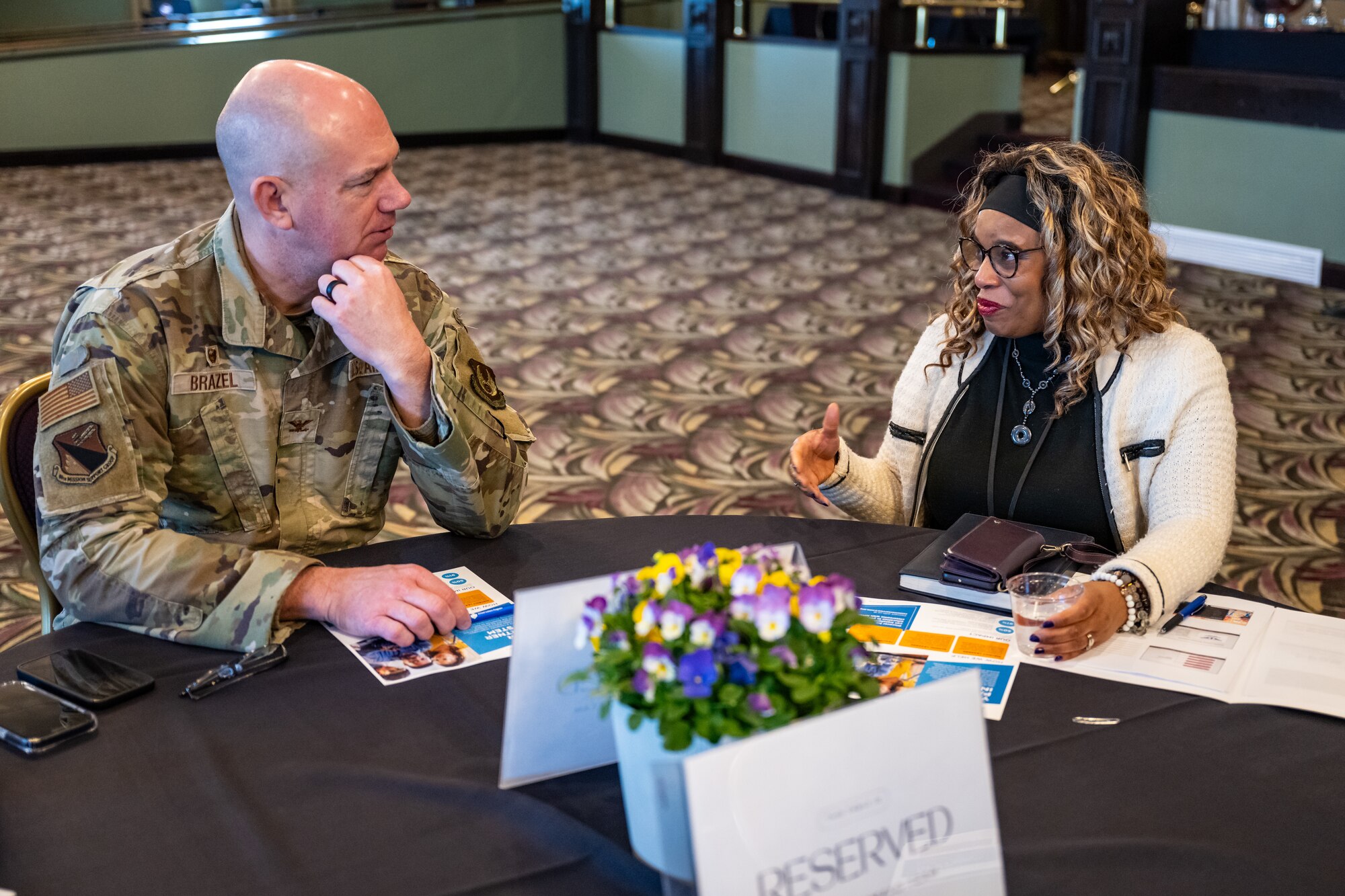 Col. Brazel, in military uniform, sits and converses with a woman at a table with documents and pot of flowers