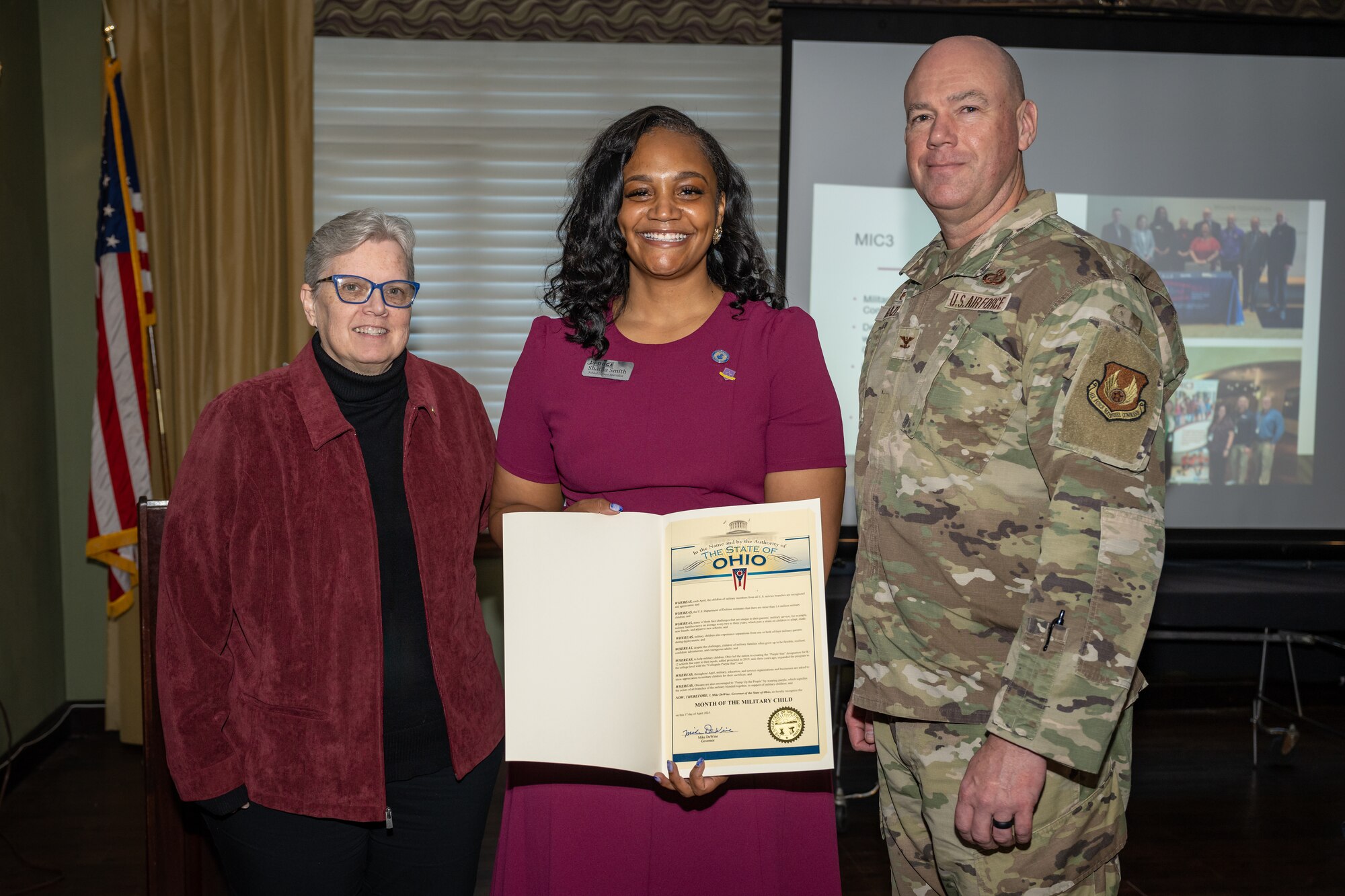Two women stand to the left of a man in military uniform. The woman in the center holds an official document