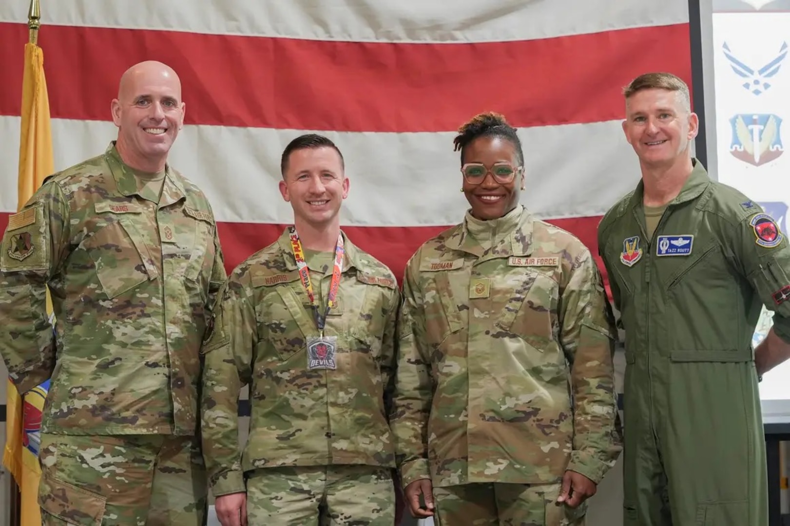 Four people in uniform stand in front of a large American flag looking at the camera.