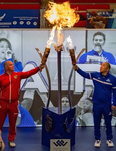 Marine at left, Airman at right holding torches toward a larger flame in the center.