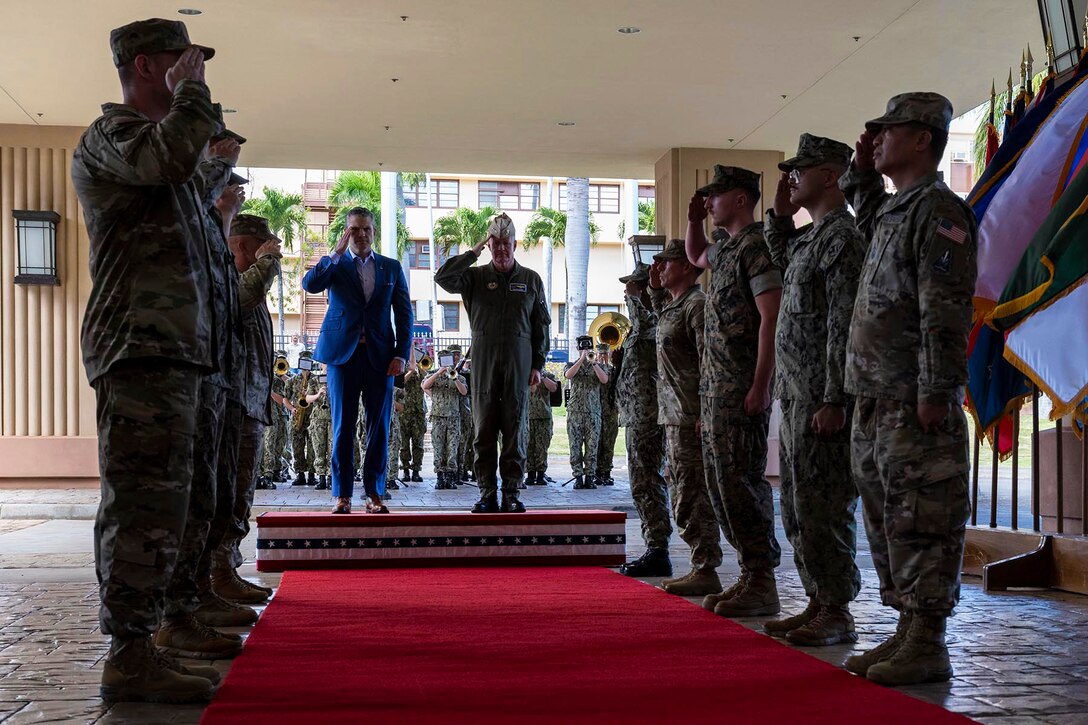 Defense Secretary Pete Hegseth and a military commander salute at the entrance to a room as saluting troops line a red carpet.