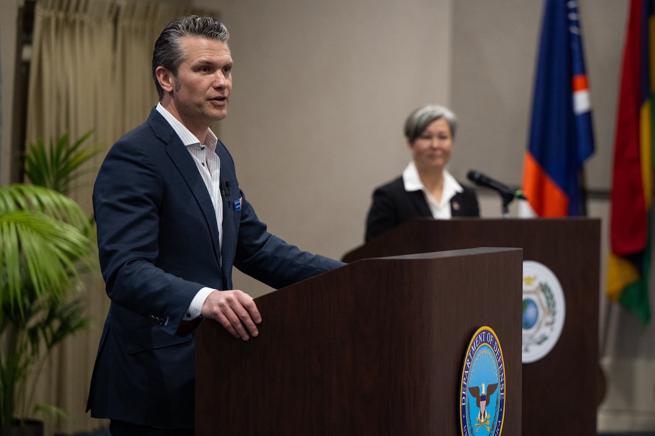 Defense Secretary Pete Hegseth speaks from behind a lectern next to another person also at a lectern with a flag and a plant in the background.