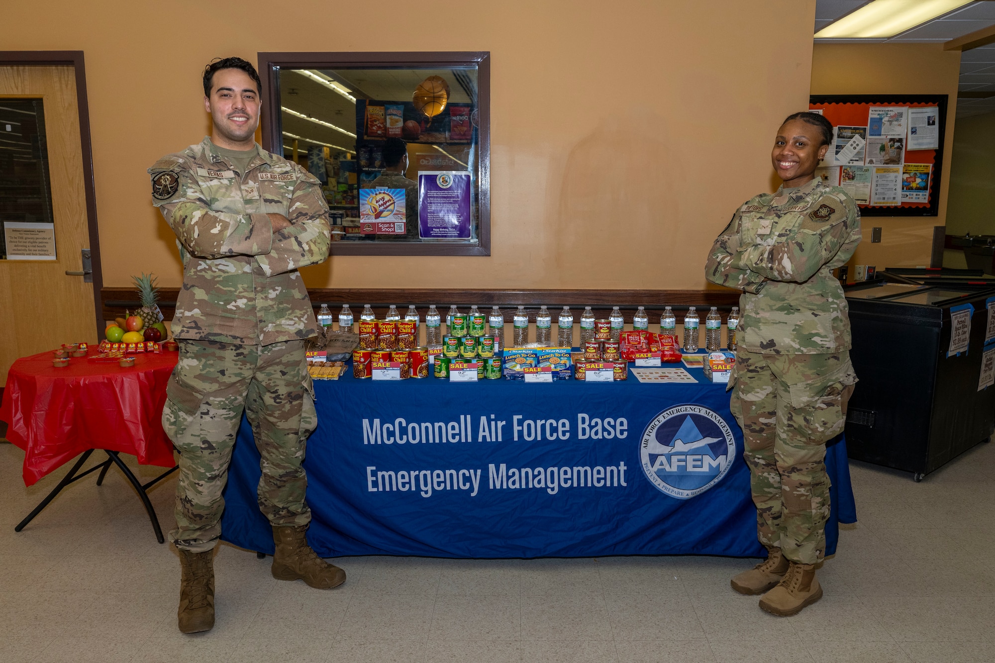 Senior Airman Danbry Veras and Airman 1st Class Ledena Baytops, 22nd Civil Engineer Squadron emergency management logisticians, pose for a photo in front of an emergency management booth at McConnell Air Force Base, Kansas, Feb. 25, 2025. The booth was set up to educate Team McConnell on the importance of food preparedness during tornado season. (U.S. Air Force photo by Airman 1st Class Paula Arce)