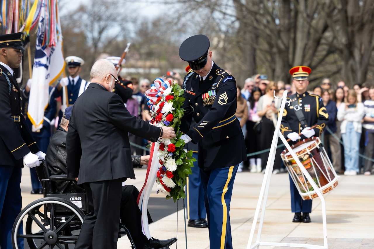 A soldier in ceremonial uniform lays a wreath while veterans look on.