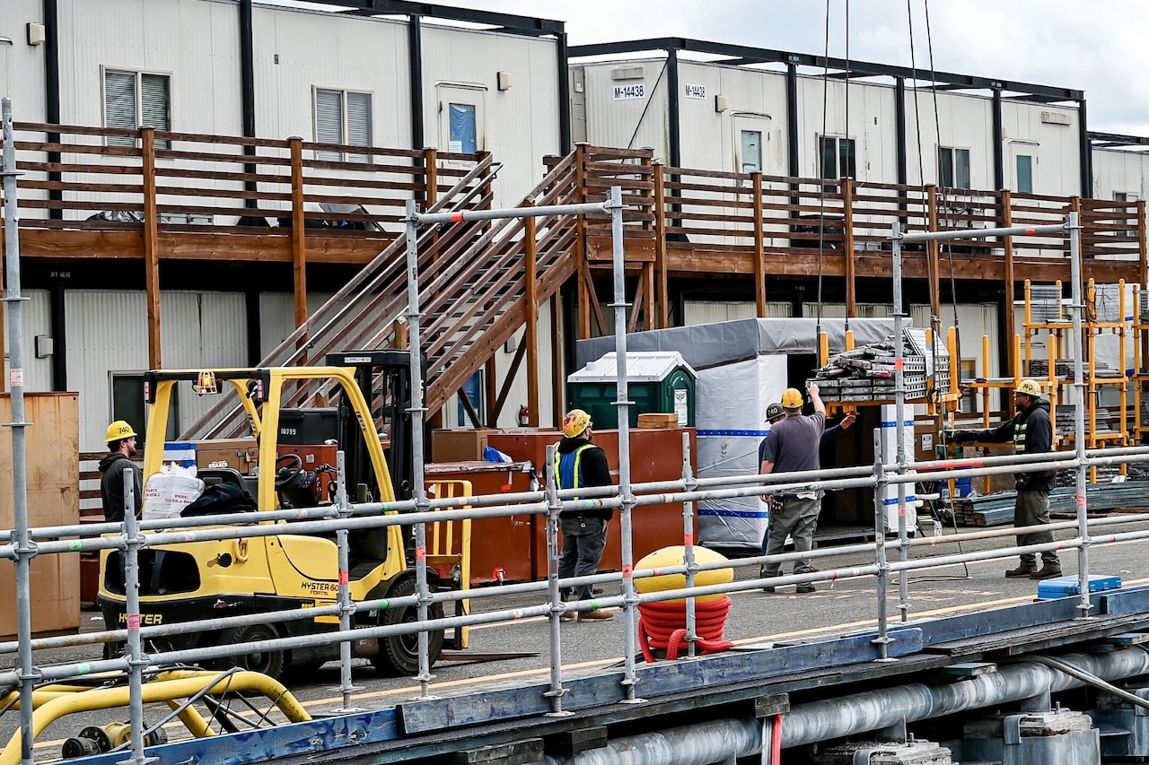 Four people work to lower items from a crane onto a pier. Another person watches from a vehicle.