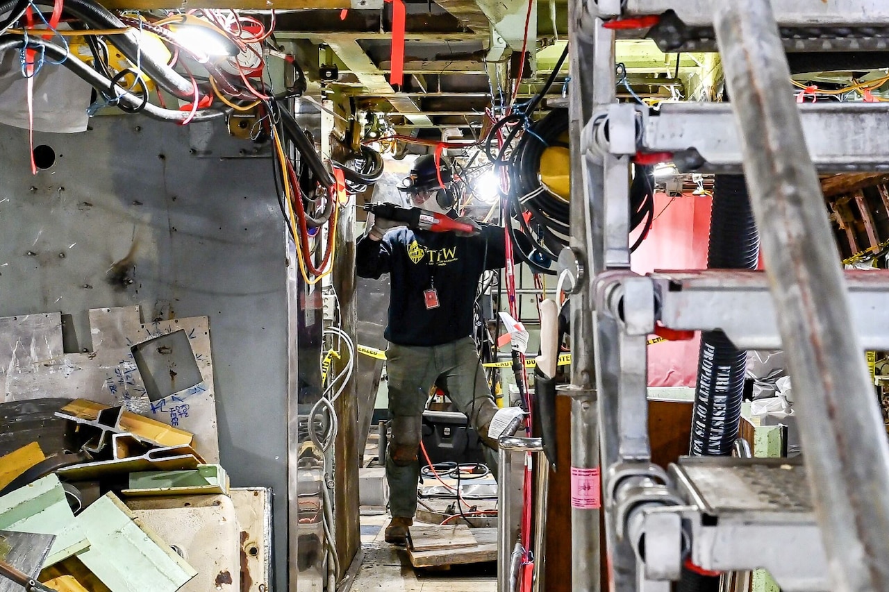 A person wearing a helmet and goggles uses a saw to cut pieces from an item in a cluttered room.
