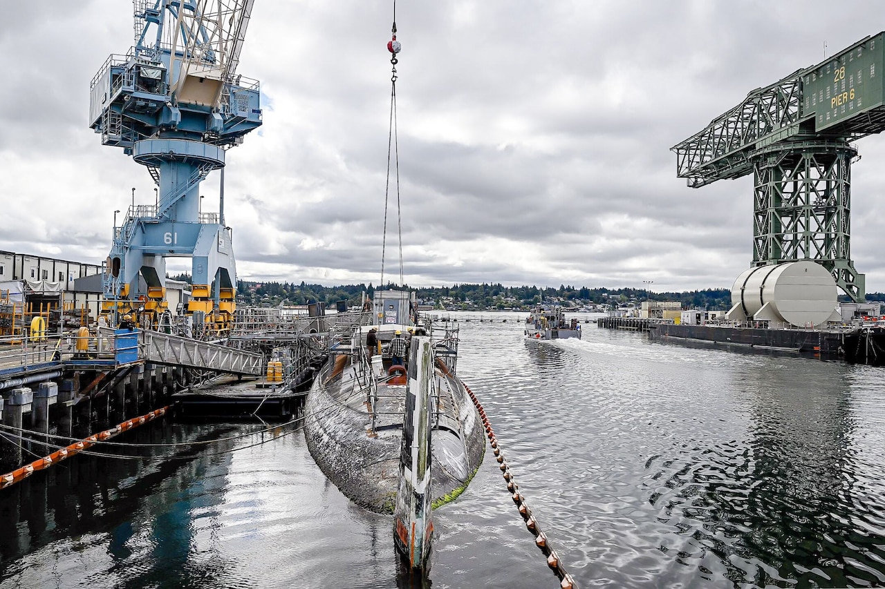 A crane on a pier sits beside a floating submarine as a small ship moves through the water in the background. A second hammerhead crane is in the background.