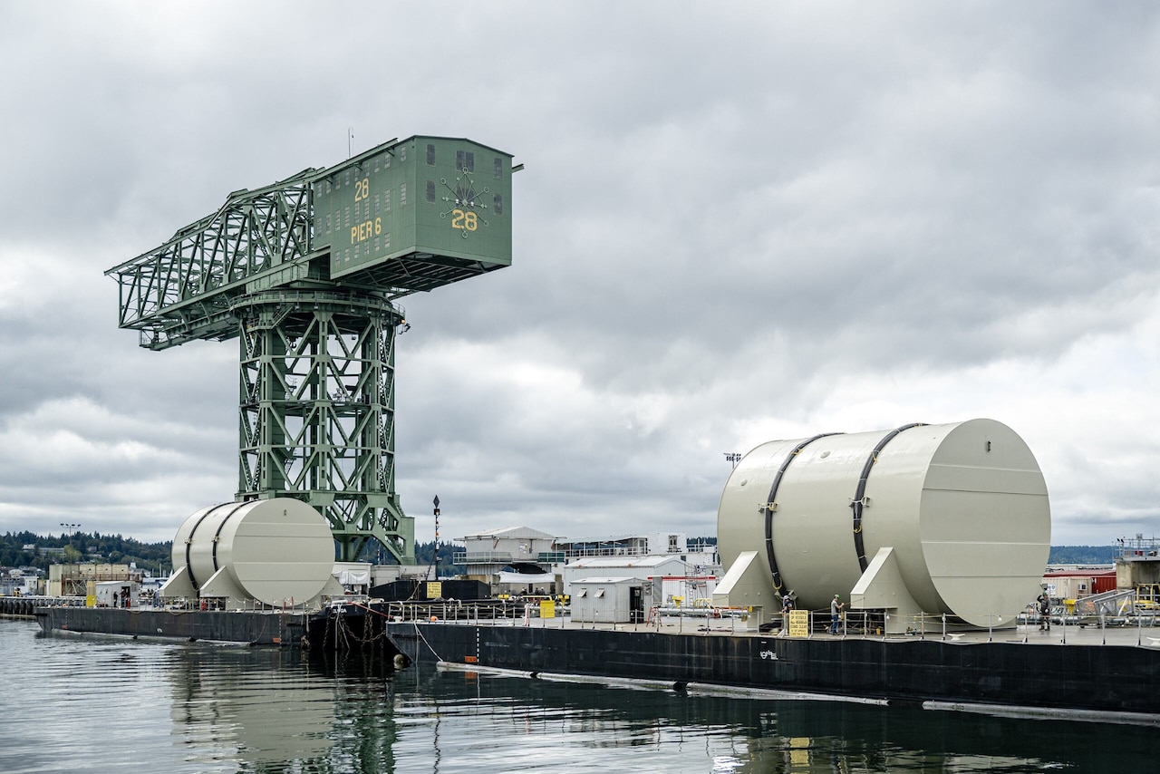 Two massive round containers sit on a pier near a large hammerhead crane.