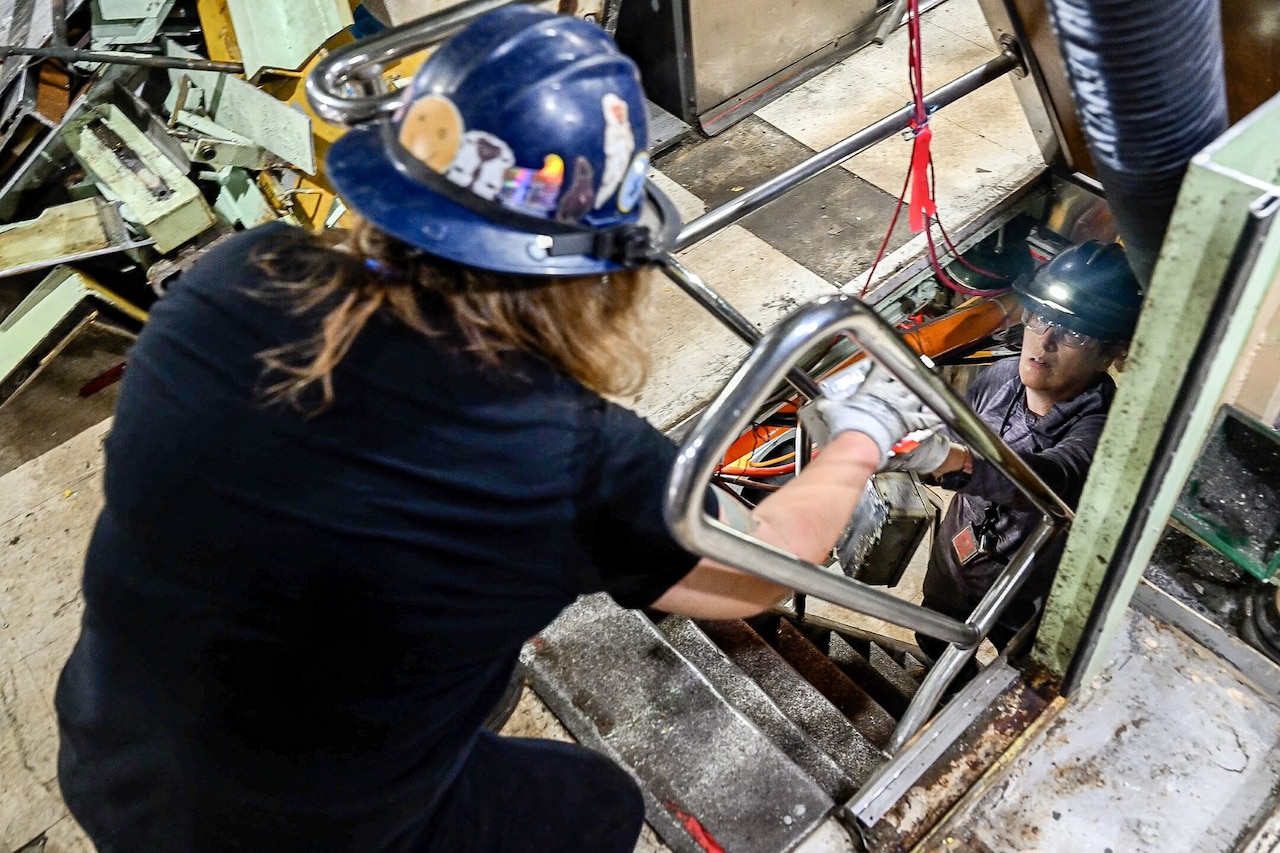 A person hands something up to another person from a narrow stairwell surrounded by debris.