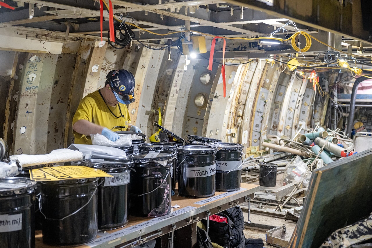 A person looks into one of several drums placed on a table inside a cluttered, dismantled submarine.