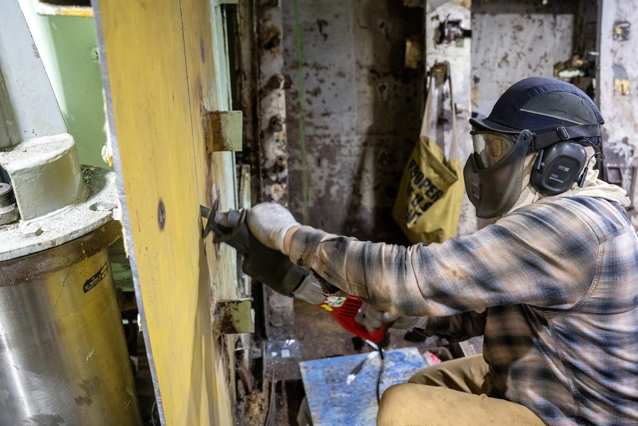 A person wearing a helmet and face mask uses a saw to cut pieces of a wall.