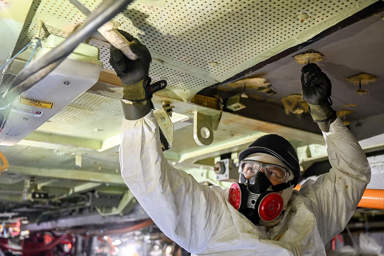 A person wearing protective equipment scrapes a ceiling.