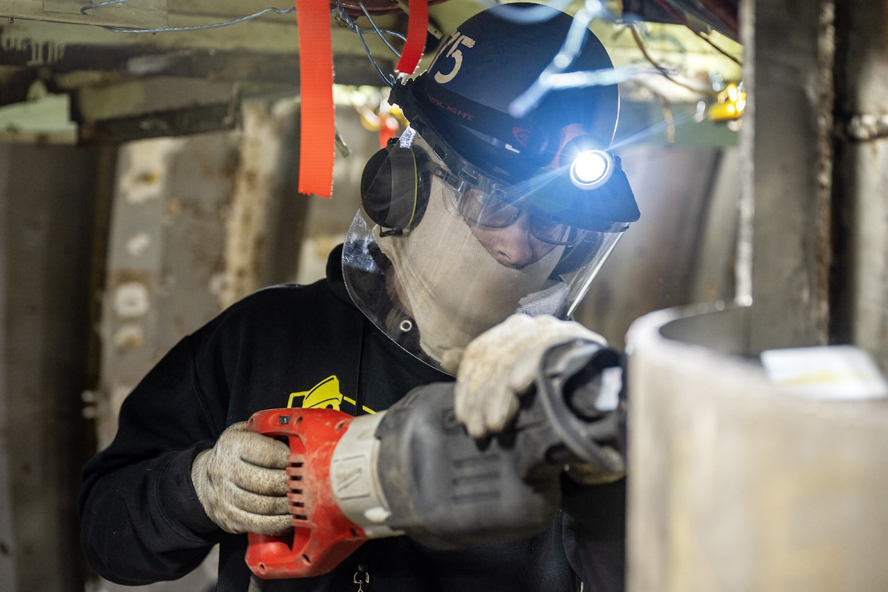A person wearing a helmet with a headlamp uses an electric saw to cut a piece of steel.