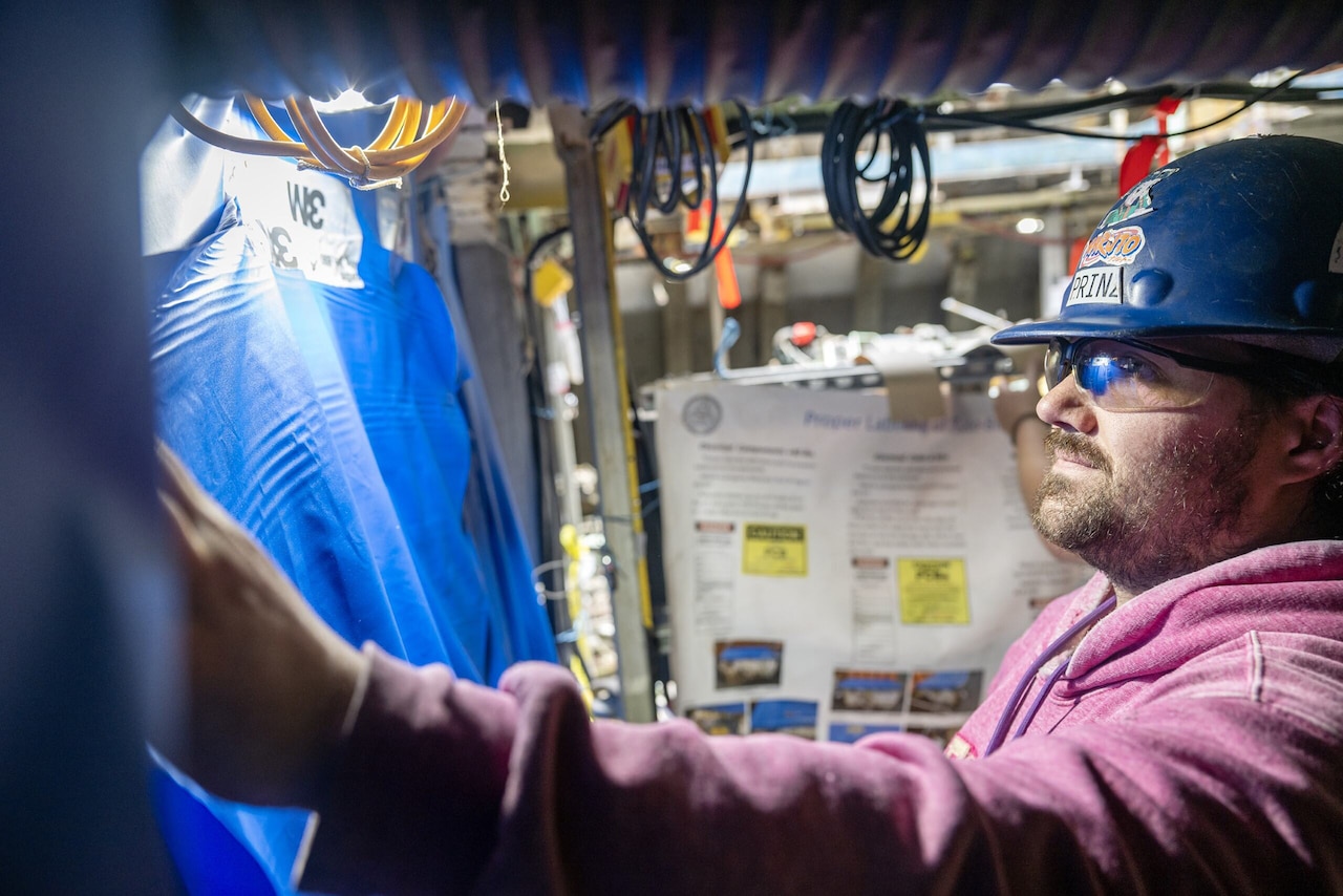 A man wearing a hard hat and goggles looks at a blue tarp.