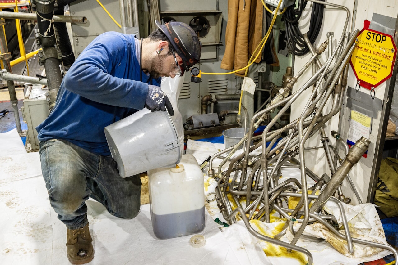 A man pours liquid into a bucket on the floor of a workspace surrounded by pipe pieces.